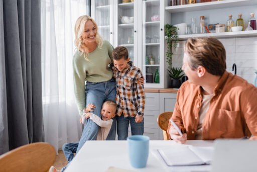 Smiling parents working from home in their kitchen, with young children embracing mom.